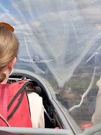 Foto aus dem Cockpit eines Segelflugzeugs im Schlepp-Flug. Links ist die Pilotin von hinten zu sehen, rechts vorne sieht man das schleppende Motorflugzeug, eine DR 401.